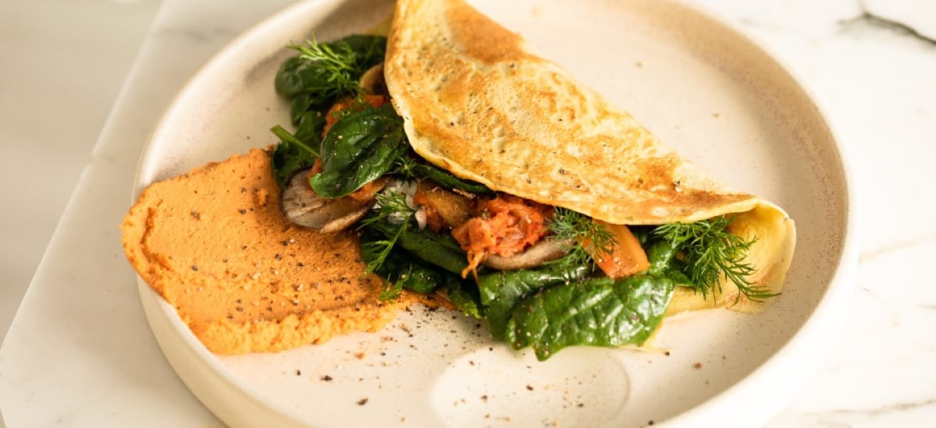 A plate with a folded omelette filled with fresh spinach, tomatoes, and herbs, accompanied by a dollop of a reddish spread on the side.